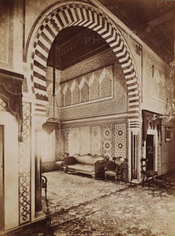 Interior of the Dar El Bey in the Medina of Tunis, Tunis, Dar-el-Bey, la Salle des Ministres, photograph, number: '367', photographer: Neurdein Frères, Medina of Tunis, 1863 - 1900, North Africa, paper, albumen print, height 273 mm x width 208 mm, palace, Dar El Bey, A richly decorated interior with a large striped archway featuring alternating light and dark bands framing a recessed area with ornate patterned walls and a carved frieze. Beneath the arch, tessellated wall panels display geometric and floral motifs. A carved wooden couch with upholstered seat and curved armrests sits against the patterned wall, flanked by matching upholstered chairs with carved legs and arms. Heavy draped curtains with fringed valances hang at openings, and a hanging chandelier with chains descends into the space. The floor is covered by an intricately patterned carpet extending across the room. Columns and pilasters with decorative inlays and carved capitals support the arch and frame doorways, and smal...