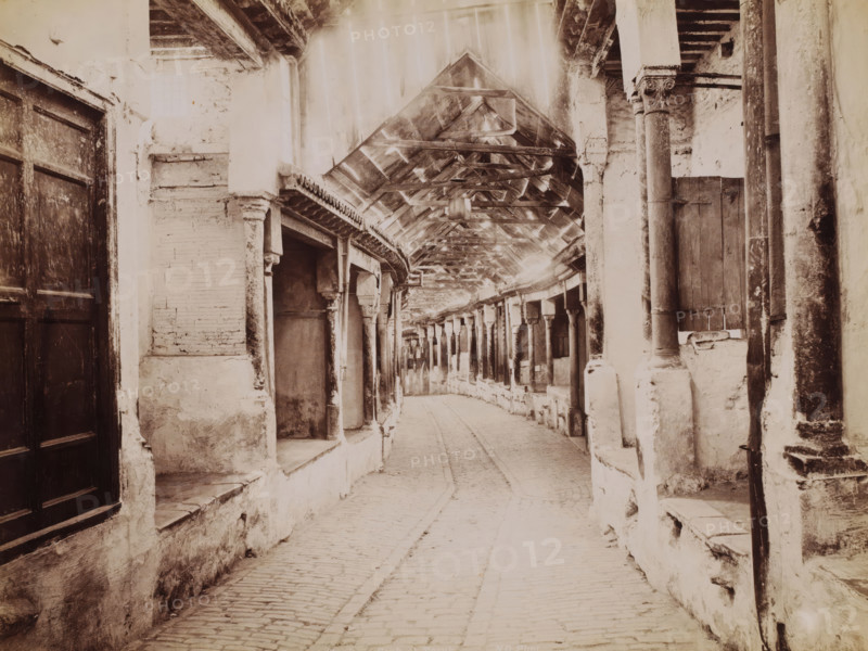 Tunis, Souk-el-Trouk, View in the Souk el-Trouk in the Medina of Tunis (abandoned), photograph, number: '287', photographer: Neurdein Frères, Medina of Tunis, 1863 - 1900, North Africa, paper, albumen print, height 205 mm x width 273 mm, market-hall, Souk el-Trouk, A narrow covered walkway with a tiled, pitched roof supported by a row of carved columns and beams runs away from the viewer, flanked by weathered plaster walls and recessed alcoves; a cobblestone path follows the center, bordered by low stone ledges and worn steps, while wooden shutters and large wooden doors are set into the walls, and patches of peeling plaster, exposed brick, and rough stone are visible along the passage.