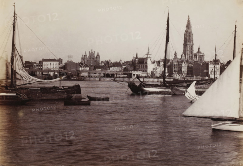 View of Antwerp from the Scheldt, Belgium, photograph, Antwerp, 1851 - 1900, photographer, paper, albumen print, height 190 mm x width 280 mm, sailing-ship, sailing-boat, city-view in general, 'veduta', parts of church exterior and annexes: tower, Scheldt, Cathedral of Our Lady (Antwerp), A waterfront scene showing several sailing vessels moored on calm water with visible hulls, masts, rigging, and partially furled sails, a small rowboat floating near the larger boats, and a large triangular sail in the foreground at the right; across the water a stone quay and a row of buildings rise with varied rooflines, towers, spires, and a prominent tall gothic-style tower dominating the skyline, all under a pale sky.