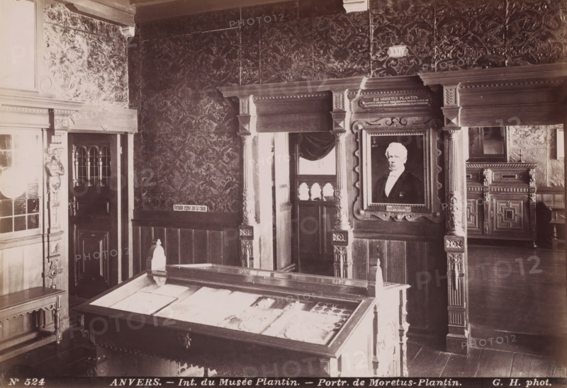 Interior of the Plantin-Moretus Museum in Antwerp, Belgium, Int. du Musée Plantin - Portr. de Moretus Plantin, Anvers, photograph, number: '76', number: 'N 524', photographer: Gustave Hermans, Antwerp, 1884 - 1914, paper, albumen print, height 192 mm x width 279 mm, permanent exhibition, museum, Museum Plantin-Moretus, A room with patterned wallpaper and carved wooden paneling, featuring ornate wooden columns and decorative moldings around doorways and wall openings, a framed portrait of a man in formal attire set within a carved frame, a glass-topped display case containing flat items, a carved wooden cabinet visible through an opening, a carved wooden bench and small table, and a small window with a draped curtain.