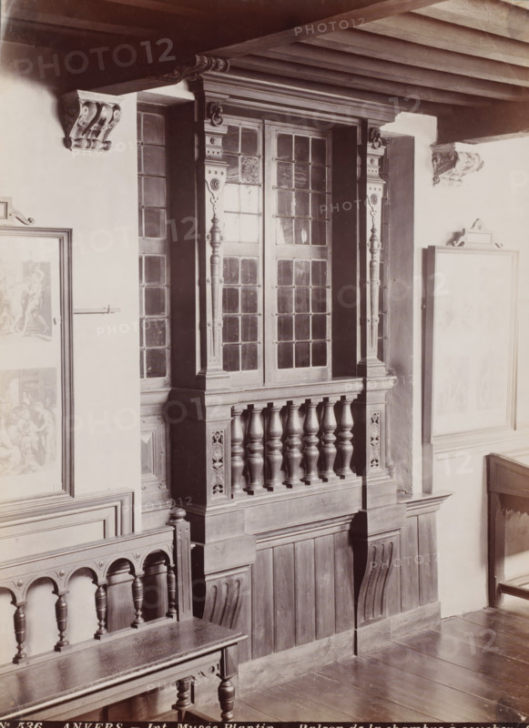 Int. Musee Plantin - Balcon de la chambre à coucher, Interior of the Plantin-Moretus Museum in Antwerp, Belgium, Anvers, photograph, number: '78', number: 'No 536', photographer: Gustave Hermans, Antwerp, 1884 - 1914, paper, albumen print, height 206 mm x width 282 mm, permanent exhibition, museum, Museum Plantin-Moretus, A sepia-toned interior view showing a wooden balcony or railing with turned balusters and carved panels beneath a set of leaded glass windows framed by decorative pilasters, fluted columns, and ornate brackets; a wooden bench with carved arches and turned legs sits in the foreground, and portions of framed pictures hang on pale walls to either side, while a beamed ceiling and wooden floorboards are also visible.