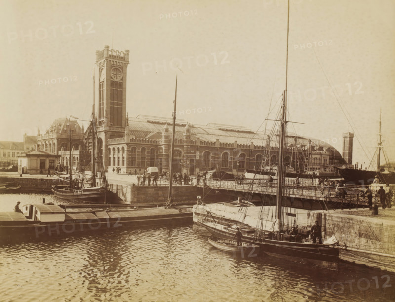 Ostende. La gare, View of the station of Ostend, Belgium, photograph, inscription: 'M&T', inscription: '... Anvers ?', number: 'No 74', Ostend, 1882 - 1900, photographer, paper, albumen print, albumendruk, height 215 mm x width 279 mm, station, shelter (railway, tramway), Ostend-City Station, A waterfront scene shows a large brick building with a tall clock tower and a long arched roof, bordered by a stone quay and a metal railing where people stand and walk; several masts, rigging, and small sailing vessels are moored along the quay, with crew or workers visible on and beside the boats, and the water in the foreground reflects ripples and shadows from the boats and structures.