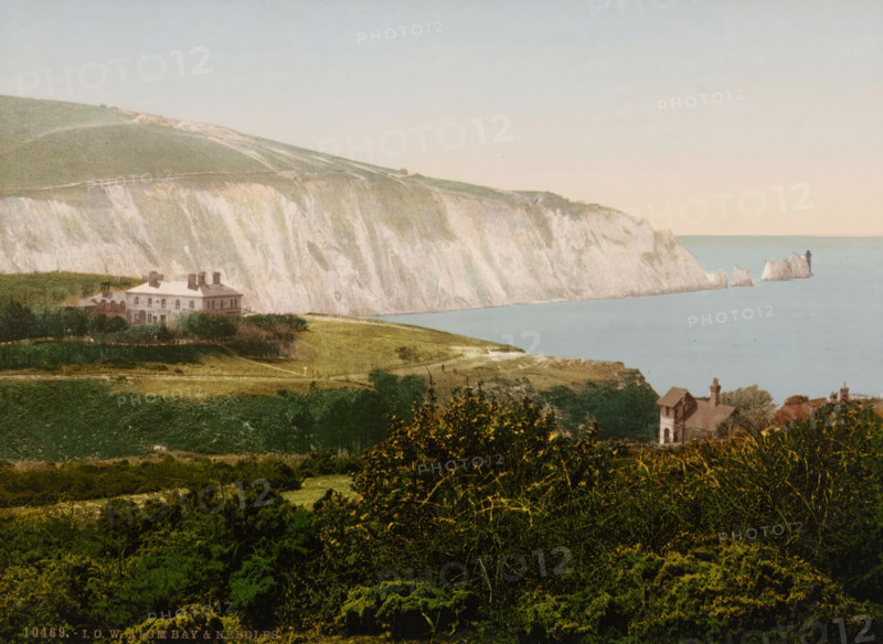 View of Alum Bay on the Isle of Wight, in the distance on the right the Needles, IOW Alum Bay & Needles, photomechanical print, number: '10469.', inscription: 'Knight's Library, 147 & 149, High St., and, Bank Buildings, Church St., Ventnor, IW', Alum Bay, cliché maker: Photochrom Zürich, Zürich (city), publisher: Photochrom Zürich, 1889 - c. 1920, Photochrom Zürich, photographer, cliché maker, publisher, Isle of Wight, paper, photochrom, height 165 mm x width 225 mm, coast, Needles, The, A coastline scene showing pale chalk cliffs rising above a calm blue sea, with a small cluster of jagged rock formations jutting from the water near the cliff edge and a dark vertical structure on one of the rocks. In front of the cliffs there are sloping grassy fields with a large pale building set back among low trees and hedges, and nearer to the foreground a smaller stone house with a pitched roof and chimney partially obscured by foliage. The foreground is filled with dense green bushes and trees,...