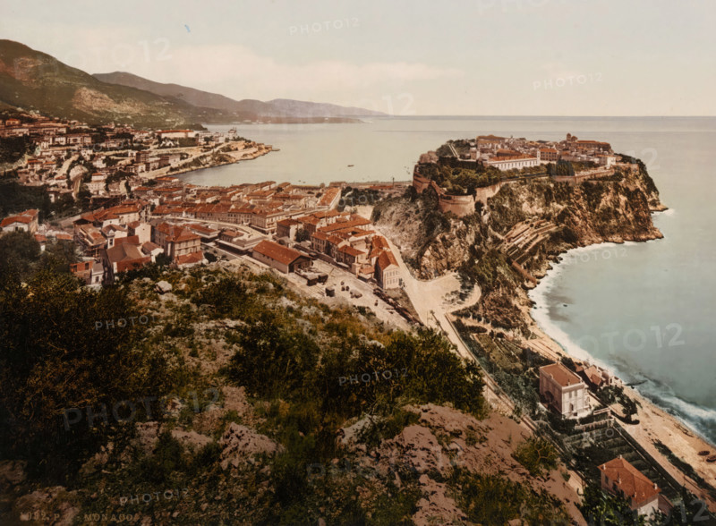 Monaco, View of Monaco, photomechanical print, number: '1082.', cliché maker: Photochrom Zürich, Zürich (city), 1889 - c. 1920, photographer, paper, photochrom, height 165 mm x width 223 mm, city-view in general, 'veduta', A coastal town seen from a high vantage point with a rocky cliffside jutting into a calm sea, a densely packed cluster of red-roofed buildings and narrow streets spreading across the slope, terraces and pathways carved into the cliff, a fortified promontory topped by larger structures and greenery, a harbor inlet with a few small boats on the water, rolling hills and more buildings in the background, vegetation and rocky ground in the foreground, and a pale sky above.