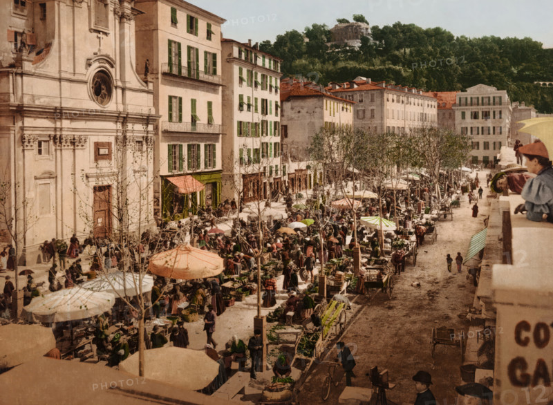 Nice, le Marché, View of the market in Nice, bottom right a photographer with camera, photomechanical print, number: '1759.', cliché maker: Photochrom Zürich, Zürich (city), 1889 - c. 1920, photographer, Provence-Alpes-Côte d'Azur, paper, photochrom, height 167 mm x width 226 mm, market, photographer at work, A wide street market scene with rows of stalls and umbrellas stretching along a tree-lined avenue beside tall pale buildings with green shutters and balconies, a large ornate building facade on the left with a round window and carved details, many people milling between carts and stalls carrying baskets and goods, vendors and shoppers gathered under patterned umbrellas and awnings, wooden carts and produce displayed on tables, leafless trees planted along the street, a hillside covered with dense trees and a stone structure visible above the treeline in the background, and a stone balustrade in the foreground where several children and a few adults lean and look down toward the ma...