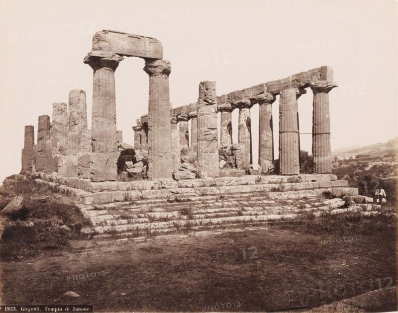 Ruin of the temple of Hera Lacinia in Agrigento, Girgenti. Tempio di Junone, photograph, number: 'No. 1833.', inscription: 'Rive (t)', photographer: attributed to Roberto Rive, Agrigento, 1860 - 1889, Sicily, Attributed to, pasteboard, paper, albumen print, height 197 mm x width 249 mm, height 307 mm x diameter 384 mm, ruin of a building, architecture, Temple of Hera Lacinia, A sepia-toned photograph of ancient stone ruins showing a row of tall, fluted columns standing on a stepped stone platform with some columns topped by large horizontal lintels and others partially broken, rough-hewn blocks and rubble around the base, sparse grass and low vegetation growing between the stone steps, an open sky above, and a person seated on a stone at the far right edge of the scene.