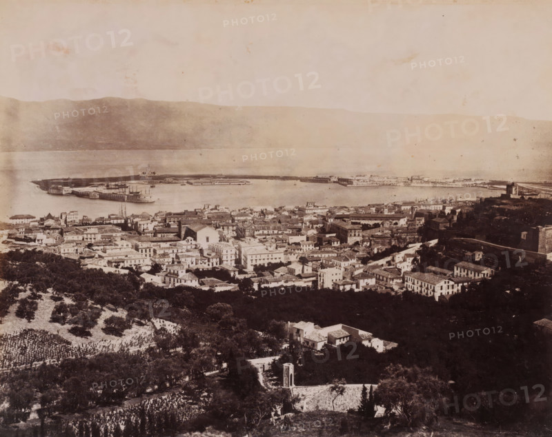 Panorama dal Casteluggio, Messina, View of Messina, Italy, photograph, inscription: 'Sommer', number: '8627', number: 'No. 1359', photographer: Giorgio Sommer, 1857 - 1914, Sicily, paper, albumen print, height 196 mm x width 241 mm, city-view in general, 'veduta', gulf, bay, A wide sepia-toned aerial view of a coastal town with densely packed buildings and tiled roofs in the foreground leading down to a harbor and a curved pier extending into calm water, with several ships anchored alongside the pier and a low mountain or hillside visible across the water in the background; scattered trees and cultivated plots appear on the slopes in the lower part of the image, and the sky above is pale and lightly mottled.