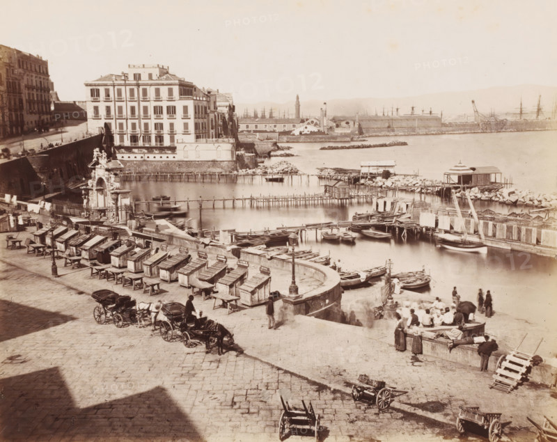 Naples rue Sainte Lucie, View of a quay in Santa Lucia in Naples, Italy, photograph, number: '12', number: '18', Naples, 1851 - 1900, photographer, pasteboard, paper, albumen print, albumendruk, height 305 mm x width 403 mm, pier, quay, wharf, A wide waterfront scene shows a stone quay with rows of wooden stalls and benches along the edge, small open boats moored in calm water, and wooden piers and platforms extending into the harbor. A multi-story pale building with many windows stands near the water beside a decorative stone gate topped with sculptural figures. People in period clothing are gathered near the quay, some sitting on the edge, others standing or walking. Horse-drawn carriages are parked on the cobbled area in the foreground. In the distance across the water are low industrial buildings, a tall narrow tower, several cranes, and masts of ships visible against a pale sky. Rocks and rubble form small breakwaters along the shoreline, and a few small shed-like structures sit o...