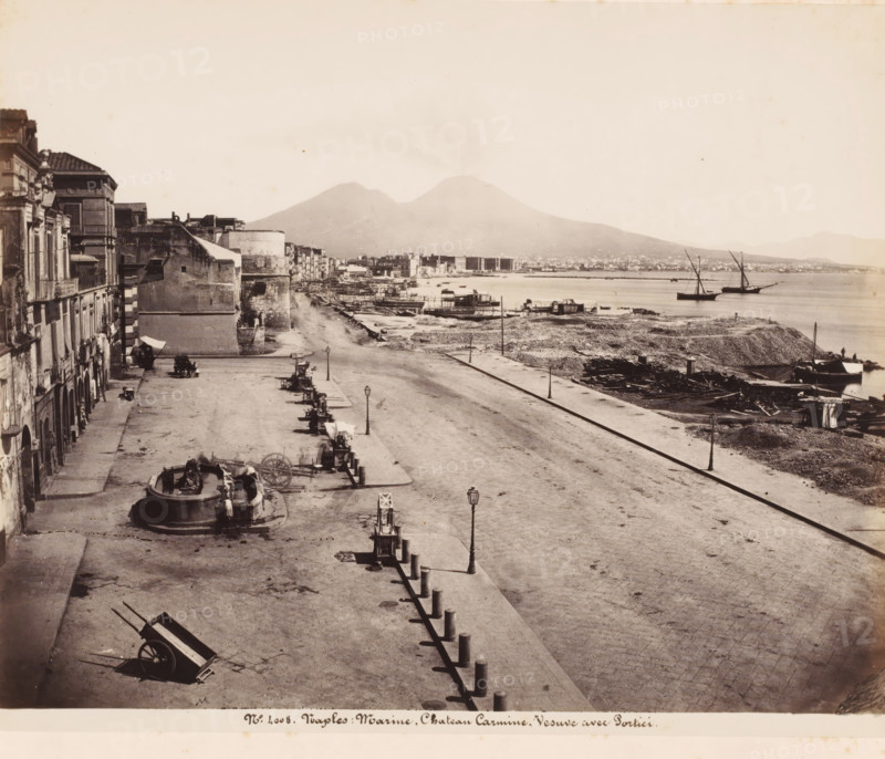 View of the harbor of Naples and Mount Vesuvius, Italy, Marine, Chateau Carmine-Vesuve avec Portici, Naples, photograph, number: '12 -', number: 'No 4008', number: '12', photographer: attributed to Giorgio Sommer, 1857 - 1914, Attributed to, pasteboard, paper, albumen print, height 307 mm x width 404 mm, harbour, volcano, A wide waterfront scene showing a broad paved promenade running alongside a shoreline with scattered debris and wooden planks, a row of short bollards lining the edge, and several lampposts along the promenade; to the left there are multi-story buildings with balconies and arched doorways, a circular stone fountain with figures, and a few handcarts and wagons positioned on the pavement; in the middle distance there are low stone structures and small waterfront platforms and docks extending into the water; on the water two sailing vessels with tall masts are anchored near the shore; in the background a large double-peaked mountain rises with a faint plume of smoke or h...