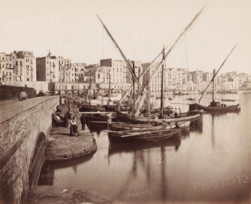 Harbour view in Naples, Italy, Naples la Marine, photograph, number: '12', number: '13.', Naples, 1851 - 1900, photographer, pasteboard, paper, albumen print, albumendruk, height 306 mm x width 404 mm, harbour, sailing-ship, sailing boat, A waterfront scene showing several wooden sailboats moored along a stone quay with tall, angled masts and rigging rising above the boats, calm reflective water, a stone wall and stepped edge along the quay with bollards and coiled ropes, a few people standing and sitting on the quay near the boats, and a long row of multi-story buildings with balconies and windows lining the opposite shore.