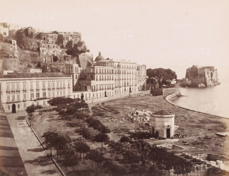 View of the Castel dell'Ovo in Naples, Italy, Chateau ... et Chateau de l'oeufs, Naples, photograph, number: '...', number: '12', number: '16 -', photographer: attributed to Giorgio Sommer, 1857 - 1914, Attributed to, pasteboard, paper, albumen print, height 307 mm x width 404 mm, urban housing, landscape with tower or castle, Castel dell'Ovo, A wide coastal scene showing a row of multi-story buildings with arched doorways and shuttered windows along a waterfront promenade, a domed roof rising among the structures, terraced stone walls and older masonry built into a hillside behind the buildings, a curved seawall and calm water beside a rocky shoreline, a square stone pavilion with an open doorway and a small roofed entrance closer to the water, scattered trees and planted areas in the foreground, a fortified stone structure on a rocky outcrop further along the coast, and various stone blocks and construction remnants visible on the ground.