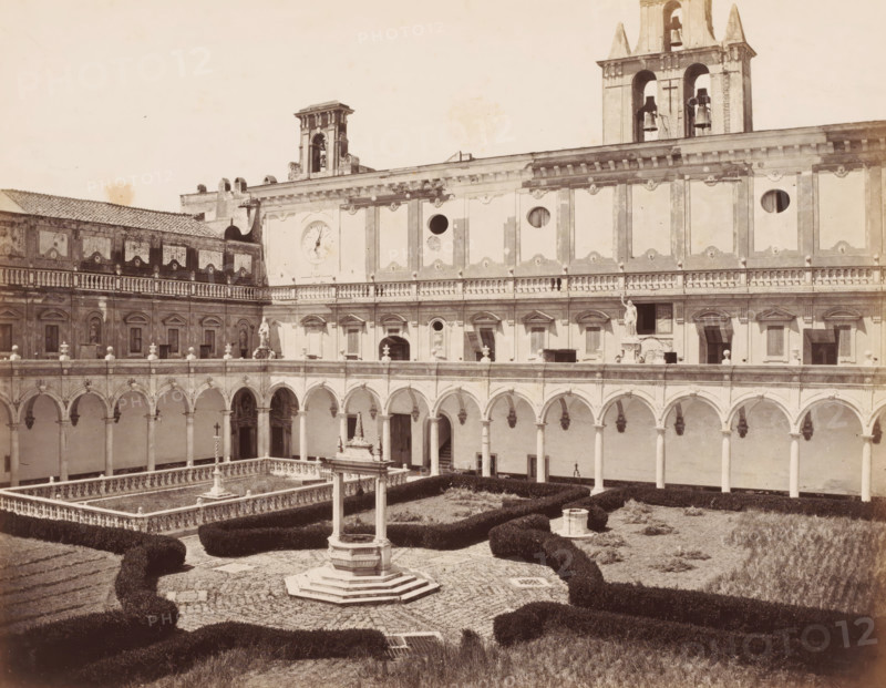 Cloitre a la Chartreuse S.Martin, Courtyard of the Charterhouse of Saint Martin in Naples, Italy, Naples, photograph, number: 'No 4070', number: '12', number: '3.', photographer: attributed to Giorgio Sommer, 1857 - 1914, Attributed to, pasteboard, paper, albumen print, height 307 mm x width 404 mm, cloisters, monastery, Carthusian Monastery San Martino, A sunlit stone courtyard with an ornate central well surrounded by a patterned paved area and low clipped hedges, framed by a two-story building with a long colonnaded arcade of rounded arches on the ground level and a balustraded gallery above, decorative rectangular wall panels and circular openings on the upper façade, sculpted statues and reliefs set into niches, a clock face on one wall, and bell towers rising above the roofline.