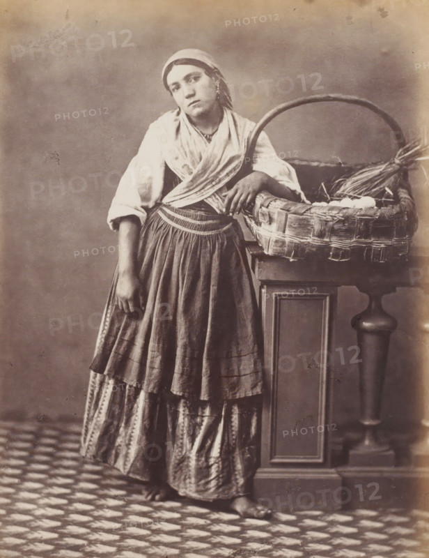 Marchande d'Oeufs, Woman with basket of eggs, Naples, photograph, number: '34', number: 'No. 4187', number: '12', inscription: 'Sommer (t)', photographer: attributed to Giorgio Sommer, 1857 - 1914, Giorgio Sommer, photographer, Attributed to, pasteboard, paper, albumen print, height 402 mm x width 305 mm, merchant, salesman, adult woman, eggs, egg dishes, A young woman stands barefoot on a patterned floor leaning against a wooden pedestal with a large woven basket resting on it, the basket containing round objects and straw; she wears a headscarf, a light shawl crossed over her shoulders, a long skirt with layered fabric and patterned lower panels, and a fitted waistband, one arm hanging by her side and the other resting on the basket rim while she gazes forward with a neutral expression.