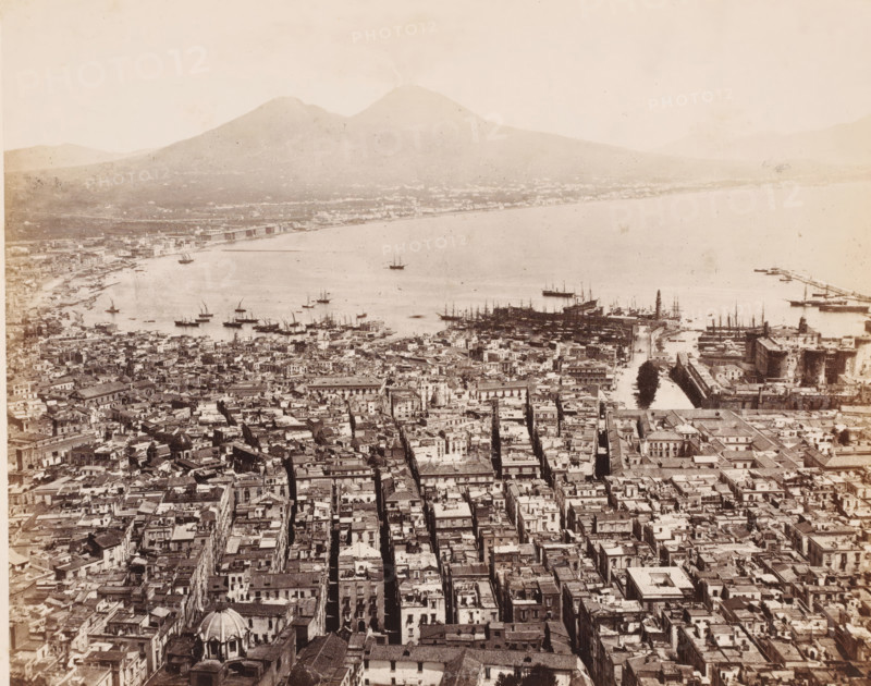 View over the Bay of Naples and Mount Vesuvius from the (former) Carthusian Monastery of Saint Martin, Italy, Panorama prize de la Chârtreuse S. Martin, Naples, photograph, number: '12', number: 'No 4003', number: '1-', photographer: attributed to Giorgio Sommer, 1857 - 1914, Attributed to, pasteboard, paper, albumen print, height 306 mm x width 405 mm, city-view in general, 'veduta', gulf, bay, volcano, Mount Vesuvius, Carthusian Monastery San Martino, A wide aerial view of a coastal city showing densely packed buildings with narrow streets running toward a waterfront lined with boats and docks, a harbor with many small and larger sailing vessels anchored and a pier extending into calm water, a prominent fortress-like structure near the water with towers and walls, a lighthouse-like tower by the docks, a distant shoreline curving around the bay, and a large mountain with a twin peak rising beyond the city under a pale sky.