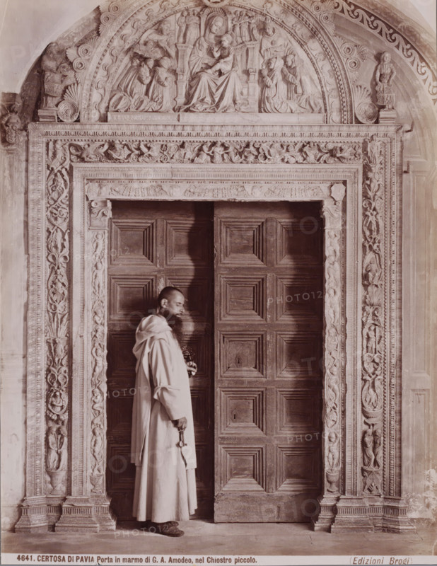 Porta in marmo di GA Amodeo, nel Chiostro piccolo, Monk standing in a marble portal in the monastery church Certosa di Pavia in Pavia, Italy, Certosa di Pavia, photograph, inscription: 'Brogi', number: '4641.', number: '13', Pavia (city), publisher: Giacomo Brogi, Florence, 1856 - 1881, photographer, Tuscany, Lombardy, pasteboard, paper, albumen print, height 404 mm x width 304 mm, monk(s), friar(s), parts of church exterior and annexes: portal, A man wearing a long robe stands in front of a large wooden door with raised rectangular panels, holding a large key in one hand and a smaller object in the other; the doorway is framed by an ornate carved stone surround with vertical decorative reliefs on either side and a horizontal carved lintel above, while an arched relief panel above the lintel depicts seated and kneeling figures carved in high relief within a semicircular arch.