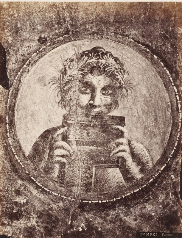 Fauno, Photo reproduction of a fresco depicting a faun, Pompei, photograph, reproduction, number: '11', number: '82.', Pompeii, after painting by anonymous, 1851 - 1900, photographer, pasteboard, paper, albumen print, albumendruk, height 400 mm x width 306 mm, picture, painting, wall-covering, satyrs, fauns, Sileni, A worn, monochrome image showing a circular framed portrait of a figure with tousled hair and laurel-like foliage woven into it, the face rendered with dark eyes and a faint smile, hands emerging from striped sleeves holding a rectangular object near the chest, surrounded by concentric circular bands and a textured, aged background with visible cracks, scratches, and spots.