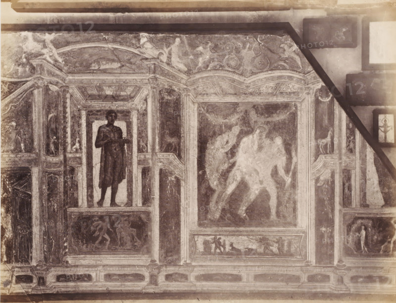 Fresco in a building in Pompeii, Italy, photograph, number: '73.', number: '11', inscription: 'Paroi Pompei', Pompeii, after painting by anonymous, 1851 - 1900, photographer, pasteboard, paper, albumen print, albumendruk, height 305 mm x width 403 mm, picture, painting, wall covering, A section of an ornate wall or ceiling paneling with framed painted scenes and decorative architectural elements, rendered in a faded monochrome tone. There are tall column-like vertical borders with carved capitals and bases separating rectangular and arched panels. One panel shows a standing male figure visible from head to feet, holding a shallow tray or platter. Another large central panel contains two overlapping human figures in motion, their forms blurred and lighter than the surrounding background. Smaller panels and friezes contain various human and animal figures in different poses, some appearing to walk or interact, and a panel shows a small animal resembling a four-legged creature on a ledge....