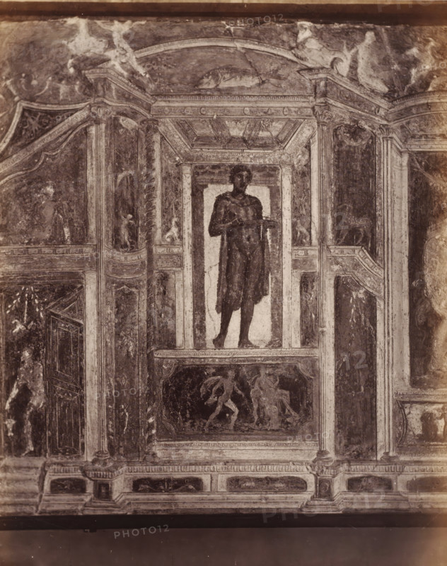 Photo reproduction of a fresco depicting a standing man in Pompeii, Italy, photograph, reproduction, number: '74.', inscription: 'Grand ... Pompei', number: '11', Pompeii, after painting by anonymous, 1851 - 1900, photographer, pasteboard, paper, albumen print, height 403 mm x width 307 mm, picture, painting, wall-covering, A faded wall painting framed by architectural painted columns and panels shows a full-length figure standing on a shallow platform within a recessed niche, the figure draped in a dark cloak and holding a shallow dish at chest height; above the niche a coffered painted ceiling and arch are visible, and below the niche a rectangular panel contains a scene of three partially rendered figures in motion; surrounding panels feature decorative motifs, small human and animal figures, fluted pilasters, and ornamental borders, all rendered in worn tones and exhibiting cracks and areas of surface loss.