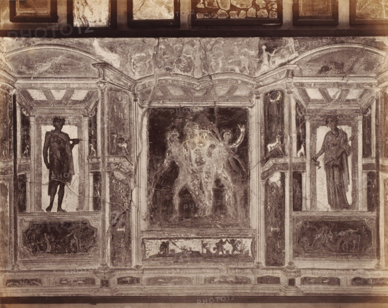 Photo reproduction of a fresco in Pompeii, Italy, photograph, reproduction, inscription: 'Parsi and bull, Pompei', number: '75.', number: '11', Pompeii, after painting by anonymous, 1851 - 1900, photographer, pasteboard, paper, albumen print, height 307 mm x width 403 mm, picture, painting, wall-covering, A faded mural panel showing architectural framing with columns and arches, flanked by two standing human figures in classical drapery within recessed niches, a central darkened scene with partially visible figures and raised arms, decorative smaller panels with animal and human motifs, ornamental moldings and rectangular compartments, and areas of surface wear, cracks, and discoloration across the painted surface.
