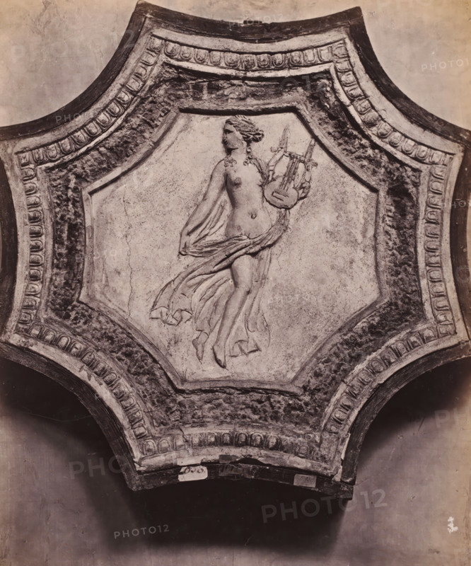 Relief depicting Medea, photograph, number: '61.', inscription: 'Stuc de Pompei, .. Medea', number: '11', Pompeii, after design by anonymous, 1851 - 1900, photographer, pasteboard, paper, albumen print, albumendruk, height 244 mm x width 199 mm, art (relief, sculpture), Medea, A carved stone panel set within an ornate geometric frame is shown from a slightly angled perspective, the panel featuring a relief of a partially draped human figure in profile holding a stringed instrument; the figure has flowing hair gathered at the back, exposed torso, and drapery that wraps around the lower body and trails downward. The surrounding frame is textured and carved with repeating rounded motifs and inset borders, showing areas of rough surface and wear. The stone surfaces display a range of tones from light to dark, with visible cracks, chips, and areas of erosion. The lower portion of the framed piece casts a soft shadow onto a plain background, and small pieces of adhesive or patched material a...