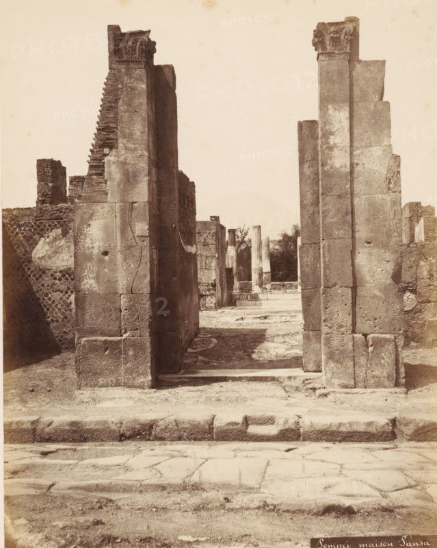 Remains of the Casa di Pansa in Pompeii, Italy, Pompei maison Pansa, photograph, number: '10', inscription: 'Pompei, maison Pansa Edile', Pompeii, 1851 - 1900, photographer, pasteboard, paper, albumen print, height 402 mm x width 304 mm, ruin of a dwelling, house, castle, Casa di Pansa, A sepia-toned photograph of a stone doorway flanked by two tall rectangular columns with carved capitals, leading into a courtyard framed by additional stone columns and ruins; the foreground shows a worn stone pavement and a low raised threshold, the walls and columns display weathered surfaces, blocks, and visible cracks, with textured masonry and scattered debris, and a narrow view through the doorway reveals more columns and fragments against a pale sky.