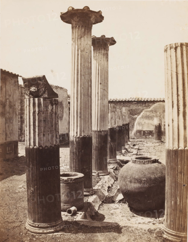 Remains of the house of Meleager in Pompeii, Italy, photograph, inscription: 'Pompei, maison Meleagre', number: '48', number: '11', Pompeii, 1851 - 1900, photographer, pasteboard, paper, albumen print, albumendruk, height 403 mm x width 305 mm, ruin of a dwelling, house, castle, A sepia-toned photograph showing a row of tall fluted stone columns with ornate capitals and weathered surfaces, some standing intact and others broken or shortened, set among low stone bases and fragments on the ground; a large rounded stone jar sits beside one column in the foreground, and a low basin and cylindrical stone drums are visible nearby; in the background there are worn plaster walls with a tiled roofline and patches of exposed masonry, and the ground is uneven with rubble and shallow trenches between the columns.