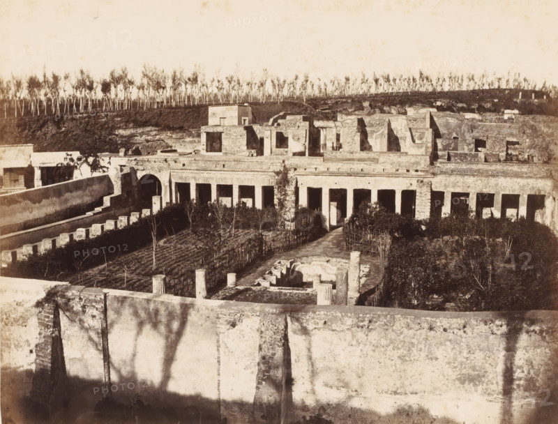 Remains of the Villa of Diomedes in Pompeii, Italy, photograph, number: '11', number: '49.', inscription: 'Pompei maison Diomede', Pompeii, 1851 - 1900, photographer, pasteboard, paper, albumen print, height 303 mm x width 402 mm, ruin of a building, architecture, A sepia-toned scene of an expansive ruined complex with stone walls, colonnades, arches, and rectangular openings, foregrounded by a high worn wall casting shadows, a central courtyard with short columns and circular stone remnants, a path leading through planted beds and trellised rows toward a colonnaded façade, and a backdrop of low stone structures and terraces topped by a line of thin, tall trees against a pale sky.