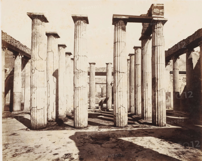 Remains of the house of Mario Epidio Sabini in Pompeii, Italy, photograph, number: '11', inscription: 'Pompei, maison Mario Epidio Sabini', number: '50', Pompeii, 1851 - 1900, photographer, pasteboard, paper, albumen print, albumendruk, height 305 mm x width 401 mm, ruin of a dwelling, house, castle, A monochrome photograph showing a row of tall fluted stone columns with square capitals standing on a paved floor, casting long shadows across the ground; some columns show surface cracks and weathering, and horizontal lintels span between a few of them; partial walls and additional columns are visible at the sides and in the background, with areas of rough stonework and shadowed recesses.