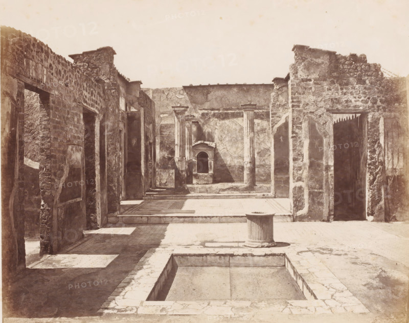 Remains of a house in Pompeii, Italy, Pompei maison Poete Tragique (Glanco) Mosaique Cave Canem fouilles 1824, photograph, number: '11', number: '25', photographer: attributed to Giorgio Sommer, Pompeii, 1857 - 1875, Attributed to, pasteboard, paper, albumen print, albumendruk, height 306 mm x width 405 mm, ruin of a building, architecture, excavation, archeology, A sunlit ruin courtyard with stone walls and columns, an empty rectangular basin set into the paved floor in the foreground, a short fluted cylindrical pedestal standing near the basin, weathered stone doorways and window openings lining both sides, textured masonry and plaster surfaces with visible cracks and patches, a shallow raised platform and mosaic-like floor area further back, and a small recessed niche framed by architectural detailing centered on the far wall.