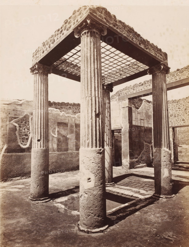Remains of a shop in Pompeii, Italy, photograph, number: '10', number: '34.', inscription: 'Pompei interior d'une boutique fouilles 1877', Pompeii, 1877 - 1900, photographer, pasteboard, paper, albumen print, albumendruk, height 307 mm x width 405 mm, ruin of a building, architecture, A sepia-toned photograph of an ancient architectural ruin showing a square canopy supported by fluted stone columns with carved capitals, a grid-like ceiling above casting a shadow pattern on the ground, surrounding low walls with patches of rough stonework and plaster, an open doorway set into a wall, and a worn earthen floor with column bases and long shadows stretching across it.