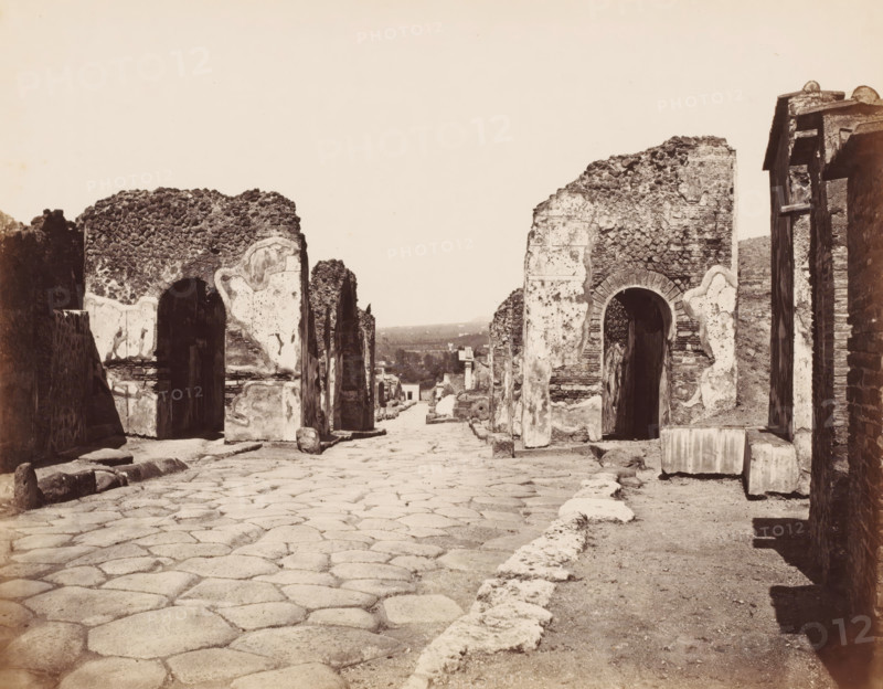 View of the remains of the gate of Herculaneum in Pompeii, Italy, photograph, number: '12.', number: '10', inscription: 'Pompei porte Herculaneum Aquelete sentinelle', Pompeii, 1851 - 1900, photographer, pasteboard, paper, albumen print, height 304 mm x width 403 mm, ruin of a building, architecture, gate, entrance, A wide stone-paved street runs toward the distance, flanked on both sides by rows of weathered stone and plaster walls with arched openings and rough, eroded surfaces, some showing exposed stonework and patches of lighter plaster; scattered stone blocks and rounded stones lie along the roadside and within openings, while the foreground shows large irregular paving stones and a raised curb edge; in the far distance low buildings and a slightly hilly landscape are visible beneath a pale, empty sky.