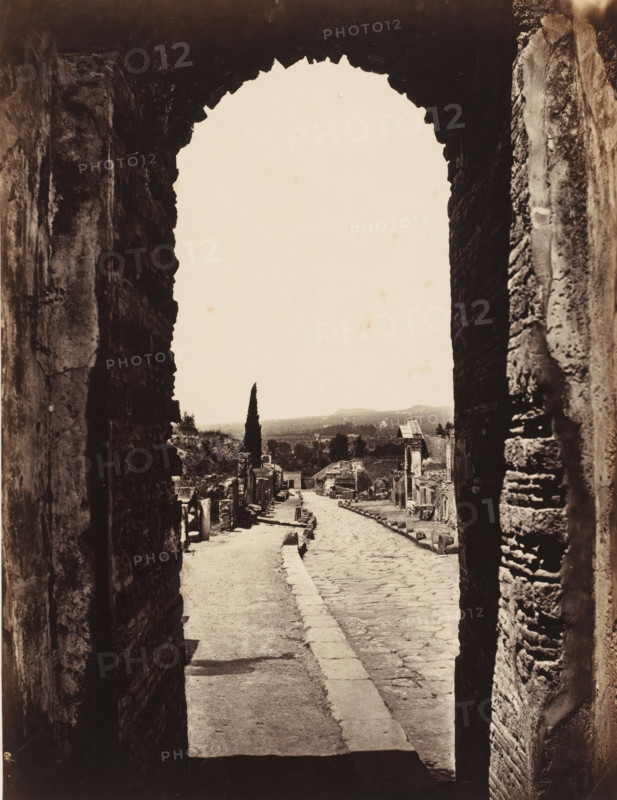 View through the gate of Herculaneum in Pompeii, Italy, photograph, inscription: 'Pompei porte Herculaneum, squelette faction', number: '20.', number: '10', Pompeii, 1851 - 1900, photographer, pasteboard, paper, albumen print, height 404 mm x width 305 mm, ruin of a building, architecture, gate, entrance, A view through a large, rough stone archway onto a narrow paved street flanked by ancient stone walls and ruins, with a cobbled path running down the center and a raised sidewalk on one side, scattered stones and fragments along the edges, a tall slender cypress tree visible in the distance against a pale sky, distant low hills on the horizon, and weathered building facades and column remnants lining the street.