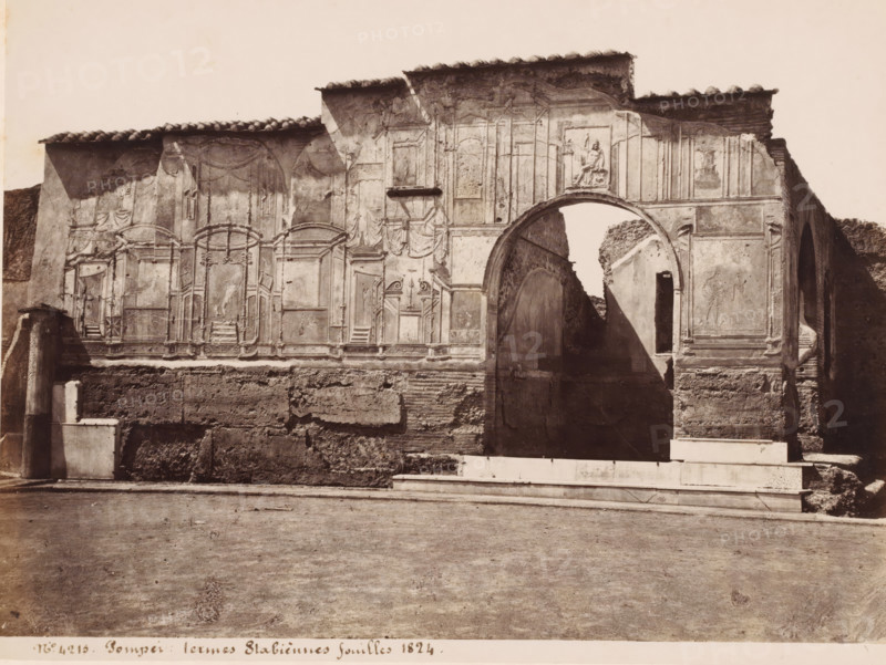 Termes Stabiennes fouilles 1824, Ruin of the Stabian Baths in Pompeii, Italy, Pompei, photograph, number: '21', number: 'No. 4215', number: '57', number: '11', photographer: attributed to Giorgio Sommer, Pompeii, 1857 - 1875, Attributed to, pasteboard, paper, albumen print, albumendruk, height 307 mm x width 405 mm, ruin of a building, architecture, excavation, archeology, A wide monochrome photograph of a weathered stone wall with architectural fresco panels and molded decorations, featuring a large rounded archway at one side revealing a narrow passage beyond and casting a diagonal shadow, a rectangular niche above the arch containing a sculpted relief of a seated figure, carved frames and drapery motifs across the wall surface, a tiled roofline with uneven eaves, rough exposed masonry at the lower portion of the wall, a stepped white stone base in front, and a flat dirt foreground extending to the bottom edge of the image.