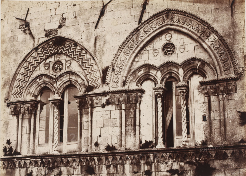 Gothic windows of the Palazzo Montalto in Syracuse, Sicily, Finestre gothique del Palazzo Montalto (Siracusa), photograph, number: 'No. 1377', number: '22', photographer: Giorgio Sommer, Syracuse (city), 1857 - 1914, pasteboard, paper, albumen print, height 312 mm x width 386 mm, open window, parts of church exterior and annexes, A close view of a weathered stone facade showing two adjacent arched window openings with ornate carved surrounds and decorative columns; the left arch features a zigzag chevron pattern and a small circular floral motif above paired narrow windows with twisted and smooth shafts, while the right arch displays a leaf and geometric border with a round tracery opening above three narrow arches and a pair of twisted columns flanking a recessed doorway; the stone surface shows darkening and mossy patches, chipped edges, and layered masonry blocks with a horizontal carved cornice and a row of triangular leaf-like decorations along the lower band.