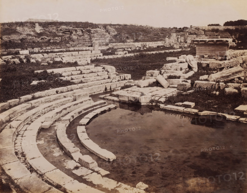 Siracuse Bagno di Diana, View of the ruins of the Bagno di Diana in Syracuse, Sicily, photograph, number: '2641', photographer: Giorgio Sommer, Syracuse (city), 1857 - 1914, paper, albumen print, albumendruk, height 199 mm x width 250 mm, landscape with ruins, A sepia-toned scene of stone ruins with curved stepped seating descending toward a shallow pool of water in the foreground, flat rectangular blocks and broken columns scattered on terraced stone platforms, low grassy areas and patches of vegetation between the masonry, a partially preserved stone structure with decorative molding on a raised platform to the right, and a low earthen bank with rough stone walls and shrubs along the back of the site under a pale sky.