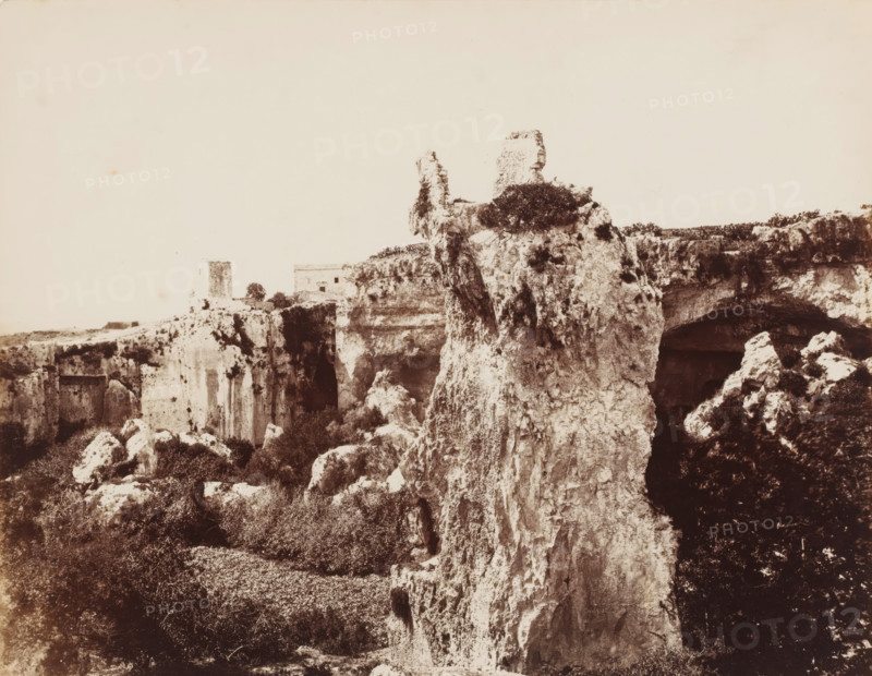 Latomie del Paradiso in Siracusa, View of Latomie del Paradiso in Syracuse, Sicily, photograph, number: 'No. 1384.', photographer: Giorgio Sommer, Syracuse (city), 1857 - 1914, pasteboard, paper, albumen print, height 309 mm x width 385 mm, quarry, A sepia-toned landscape showing a rugged cliff face with rough, weathered rock formations rising prominently in the center and right, textured with crevices and patches of vegetation clinging to ledges. In the midground a tall, narrow pillar of rock stands apart from the main cliff, topped with low shrubs, and a flat-topped ruin sits further back on the plateau. Dark cave-like openings and overhangs are visible along the cliff line, while scattered boulders and dense low bushes occupy the foreground, creating a contrast between the pale limestone surfaces and the darker foliage. The sky above is pale and largely empty.