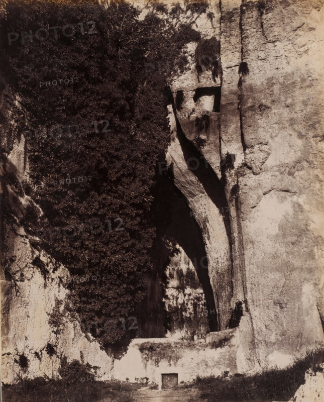 View of the Ear of Dionysius in Syracuse, Sicily, Orecchio di Dionisio, Siracusa, photograph, number: '2664', photographer: Giorgio Sommer, Syracuse (city), 1857 - 1914, pasteboard, paper, albumen print, height 254 mm x width 202 mm, cave, grotto, Ear of Dionysius, A tall vertical rock face fills the scene with rough textured stone on one side and a dark shadowed crevice on the other, a large overhanging ledge creating deep contrast where the rock curves inward; dense foliage clings to one portion of the cliff, forming a dark mass of leaves and branches against the lighter stone; beneath the overhang a small rectangular doorway set into the rock is visible with a flat area of ground leading up to it and low vegetation along the base of the cliff.