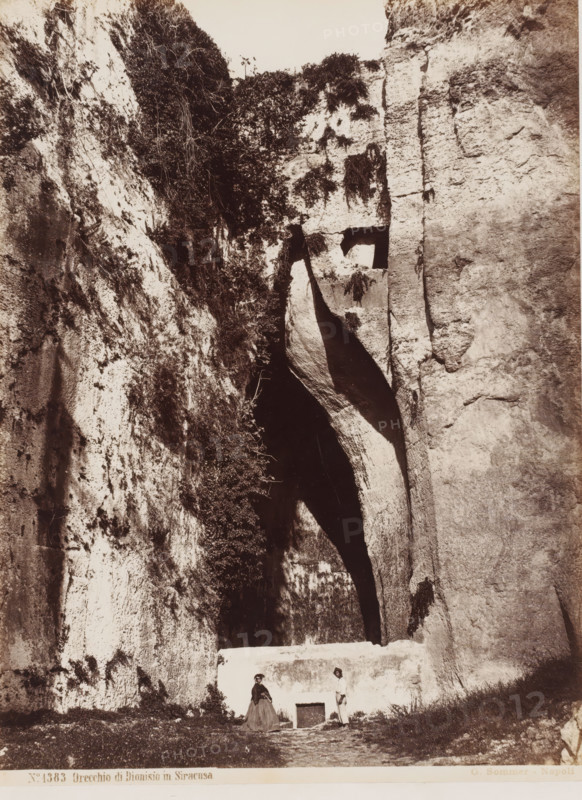 View of the Ear of Dionysius in Syracuse, Sicily, Orecchio di Dionisio in Siracusa, photograph, number: 'No. 1383', photographer: Giorgio Sommer, Syracuse (city), 1857 - 1914, pasteboard, paper, albumen print, height 384 mm x width 308 mm, cave, grotto, mountains (landscape with figures, staffage), Ear of Dionysius, A vertical rock face with rough textured surfaces fills the frame, showing lighter and darker stone areas and patches of vegetation clinging to crevices; a deep, dark archway or cave opening is set within the rock, with irregular shadows and a curved inward slope, and a flat pale ledge or wall crosses the lower part of the opening; in front of the ledge two small human figures stand on grassy ground, one wearing a long skirt and a jacket and the other wearing lighter clothing and trousers, both positioned near a rectangular dark recessed opening at the base of the ledge.
