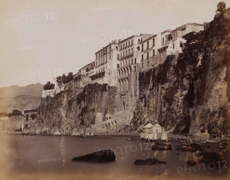 View of the cliffs of Sorrento and Hotel Tramontano, Italy, Hôtel Tramontano, Sorrento, photograph, inscription: 'Sorrento Hotel Tramontano', number: '2127', photographer: Giorgio Sommer, 1857 - 1914, paper, albumen print, height 203 mm x width 254 mm, gulf, bay, coastal cliff or crag, A sepia-toned photograph showing a dramatic cliffside with multi-story buildings perched along its edge, several of the buildings featuring balconies and arched openings; vegetation and shrubs grow on parts of the rocky face, and a series of steps and arched masonry are built into the cliff; at the waterline there are small wooden pavilions or huts on stilts connected by narrow walkways, and large dark rocks protrude from the calm water in the foreground; distant hills are visible on the left and the sky above is pale and mostly empty.