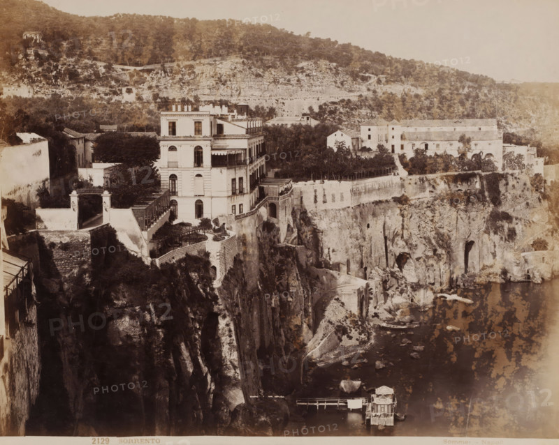 Sorrento, View of the cliffs of Sorrento, Italy, photograph, number: '2129', photographer: Giorgio Sommer, Naples, 1857 - 1914, paper, albumen print, albumendruk, height 201 mm x width 253 mm, city view in general, 'veduta', coastal cliff or crag, A wide sepia-toned view shows a large multi-story building with balconies and arched windows perched atop a rocky cliff, surrounded by terraces, stone walls, and vegetation; adjacent structures and courtyards extend along the cliff edge toward a cluster of lighter-colored buildings set back among trees; steep, jagged rock faces descend to a narrow shoreline with scattered boulders and a small wooden pier supporting a modest boathouse; paths and stairways are visible cut into the cliff and connecting levels, while the distant hillside rises with dense tree cover and scattered small buildings.