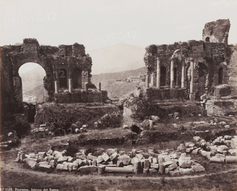 Interno del Teatro, Remains of the Amphitheater of Taormina, Italy, Taormina, photograph, number: 'No. 1745.', 1851 - 1900, photographer, Sicily, pasteboard, paper, albumen print, height 310 mm x width 384 mm, ruin of a building, architecture, amphitheatre, A wide sepia photograph shows ancient stone ruins with large arched openings and sections of crumbled walls, some topped with short columns and scattered column drums lying on the ground; the foreground contains a pile of irregularly shaped stones and broken architectural fragments on grassy earth, while midground terraces and raised platforms support remnants of masonry and upright columns; distant hills and a faint mountain silhouette appear through an archway and between the ruins, and a small cluster of buildings sits on a distant ridge; vegetation including low bushes and patches of grass grows among the ruins, and the sky above is pale and clear.