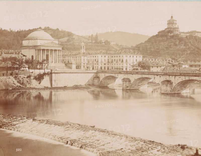 Bridge over the Po at Turin, Italy, Ponte sul Po, Torino, photograph, inscription: 'Turin, the bridge over the Po.', number: '920.', Turin, 1851 - 1900, photographer, Piedmont, pasteboard, paper, albumen print, height 257 mm x width 338 mm, bridge in city across river, canal, Po, A riverscape with a stone bridge featuring multiple arches spanning calm reflective water, a stepped weir in the foreground, a domed neoclassical building with columns near the riverbank, a row of multi-story buildings with uniform facades and tiled roofs beside it, a bell tower rising behind the row, trees and vegetation along the bank and hills in the background, and a hilltop building with a layered tower overlooking the scene.