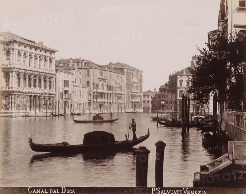 Canal dal Duca, Gondolas in the Grand Canal in Venice, photograph, inscription: 'Venetié', photographer: Paolo Salviati, Venice, 1860 - 1870, pasteboard, paper, albumen print, albumendruk, height 197 mm x width 249 mm, height 276 mm x width 363 mm, canal, gondola, Grand Canal, A wide canal scene with ornate multi-story buildings lining the far bank, featuring arched windows and decorative facades, boats and gondolas floating on the water, a gondolier standing and rowing near the center foreground, another gondola with a covered seating area closer to the front, mooring posts rising from the water along the right side, a tree partially obscuring the buildings on the right bank, stone steps and a small waterfront railing visible at the lower right edge, and gentle reflections of the boats and buildings on the canal surface.