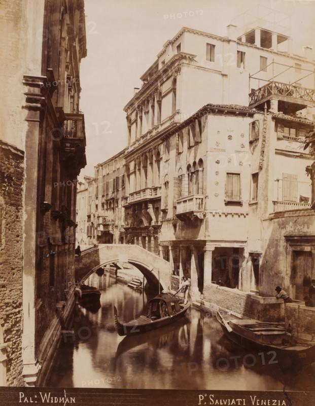 View of the Palazzo Rezzonico-Widmann in Venice, Pal. Widman, photograph, inscription: 'Venetie', photographer: Paolo Salviati, Venice, 1860 - 1870, pasteboard, paper, albumen print, height 250 mm x width 197 mm, height 348 mm x width 275 mm, urban housing, canals, waters (in city), bridge in city across river, canal, A narrow canal flanked by tall, weathered buildings with arched windows and small balconies runs under a stone bridge, with a covered gondola and other small boats on the calm water and a gondolier standing in one boat, while people sit or stand along the canal edge near columned porticos and shuttered windows, and the facades show ornamental stonework, worn plaster, and rooftop terraces.