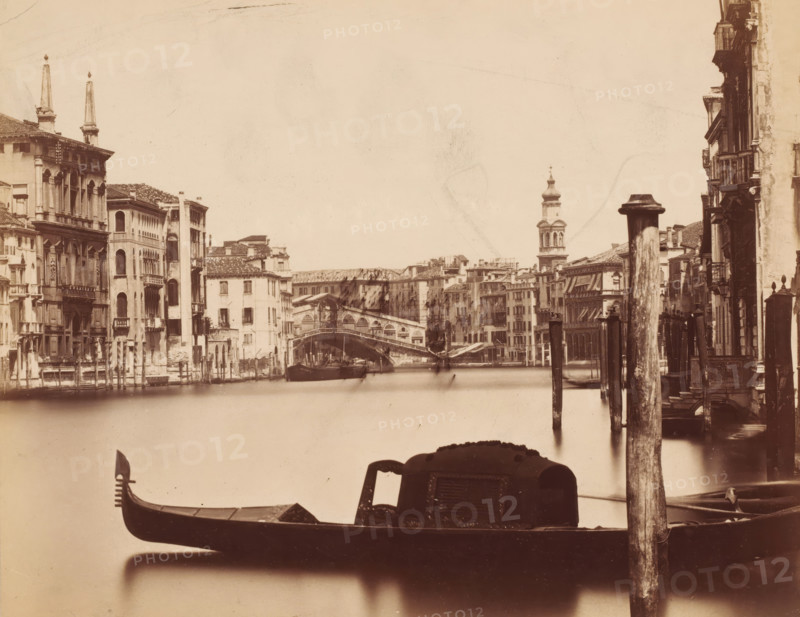 View of the Grand Canal with the Rialto Bridge in the background in Venice, Italy, photograph, Venice, 1851 - 1900, photographer, pasteboard, paper, albumen print, height 187 mm x width 240 mm, urban housing, canals, waters (in city), bridge in city across river, canal, Grand Canal, Rialto Bridge, A sepia-toned view of a canal framed by rows of multi-story buildings with arched windows and balconies on both sides, a stone bridge spanning the canal in the middle distance, a domed bell tower rising beyond the bridge, wooden posts and mooring poles along the water's edge, and a gondola with an ornate prow and covered passenger cabin in the foreground.