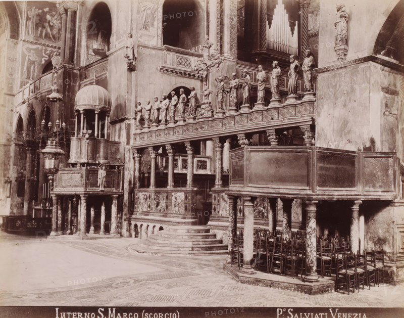 Interior of the San Marco in Venice, Interno S. Marco (scorgio), photograph, inscription: 'Venetie', photographer: Paolo Salviati, Venice, 1860 - 1870, pasteboard, paper, albumen print, albumendruk, height 197 mm x width 250 mm, height 275 mm x width 365 mm, parts of church interior, St. Mark's Basilica, A stone interior with ornate carved railings and a raised platform accessed by semicircular steps, supported by columns with decorative capitals and marbleized shafts, a row of statues standing on pedestals along the balustrade, an elevated domed pulpit with slender columns and a sculpted figure beneath it, hanging lamps and a large freestanding decorative lampstand, multiple wall niches and arches, an organ case with visible vertical pipes recessed into the wall, carved reliefs and patterned panels on the lower fronts, clustered wooden chairs lined beneath the gallery, mosaic flooring with geometric patterns, and tall wall-mounted statues and sculptural details on the surrounding surf...