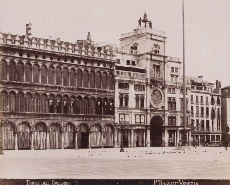 Torre dell Orologio, St. Mark's Square with the Torre dell'Orologio in Venice, photograph, inscription: 'Venetie', photographer: Paolo Salviati, Venice, 1860 - 1870, pasteboard, paper, albumen print, albumendruk, height 197 mm x width 242 mm, height 276 mm x width 365 mm, square, circus, tower, clock tower, monumental adornment, facade (of house or building), A wide view of a historic multi story stone building with rows of arched windows and shuttered openings, ornate cornices and decorative balustrades, a large central archway topped by a circular ornate clock face set into a façade with sculptural relief and a winged lion relief on an upper section, a rooftop platform with two dark human-shaped statues flanking a large bell, striped fabric awnings and curtains hanging over the lower arches, a tall slender pole rising beside the building, a paved open square in front scattered with birds and a few lampposts visible near the entrance.