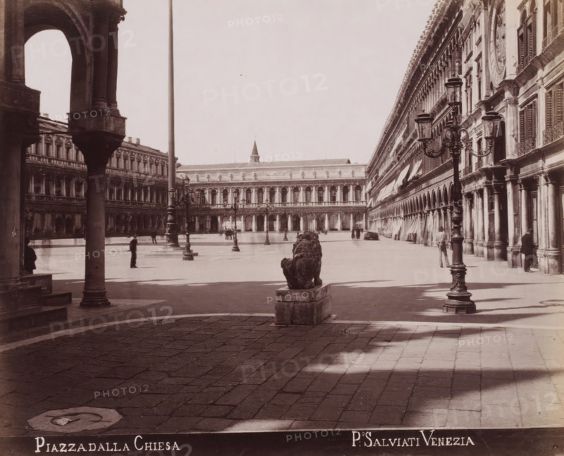 Piazza dalla Chiesa, View of St. Mark's Square from the Piazzetta dei Leoncini in Venice, photograph, inscription: 'Venetie', photographer: Paolo Salviati, Venice, 1860 - 1870, Paolo Salviati, photographer, pasteboard, paper, albumen print, albumendruk, height 199 mm x width 244 mm, height 276 mm x width 365 mm, square, circus, façade (or house or building), St. Mark's Square, Piazzetta dei Leoncini, A wide stone plaza framed by ornate arched buildings on either side with rows of columns and shuttered windows, an elevated colonnade visible across the open space, a stone lion statue on a plinth facing away from the viewer near the center foreground, tall decorative lampposts with ornate metalwork standing along the plaza, scattered figures standing or walking, shadows cast on the paved ground, and architectural details such as balustrades, cornices, and awnings along the building facades.
