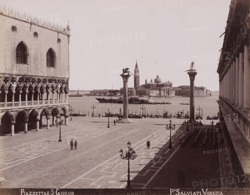 Piazzettae S. Giorgio, View of the square of the Chiesa di San Giorgio Maggiore towards the campanile of San Marco in Venice, photograph, inscription: 'Venetie', photographer: Paolo Salviati, Venice, 1860 - 1870, pasteboard, paper, albumen print, height 196 mm x width 251 mm, height 275 mm x width 364 mm, city-view in general, 'veduta', square, circus, Isola di San Giorgio Maggiore, Chiesa di San Giorgio Maggiore, A wide stone promenade bordered by ornate buildings opens toward a body of water where boats are moored and a domed church and campanile rise on the opposite shore; two tall columns topped with statues stand near the waterfront between the square and the water; on the left a palatial façade shows arched colonnades, decorative windows, and patterned stonework while on the right a long arcade of columns casts a deep shadow over rows of empty chairs and tables; ornate street lamps line the paved square where a few pedestrians are walking and small groups stand near the water’s e...
