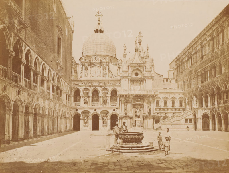 Courtyard of the Doge's Palace in Venice, Italy, Cortile Palazzo Ducale, photograph, number: 'N 25', inscription: 'Venice', inscription: 'courtyard ducal palace', Venice, 1851 - 1900, photographer, pasteboard, paper, albumen print, height 310 mm x width 400 mm, ornamental fountain, inner court of palace or 'hôtel', Doge's Palace, A wide courtyard framed by ornate stone arcades on both sides with multiple arched openings and columns, a central ornate fountain with sculpted basin where two men stand nearby, a large domed building with decorative statues and carved facades in the background, several figures including children walking or standing near the fountain and along the open paving, and a grand staircase and additional sculptural details on the building to the right.